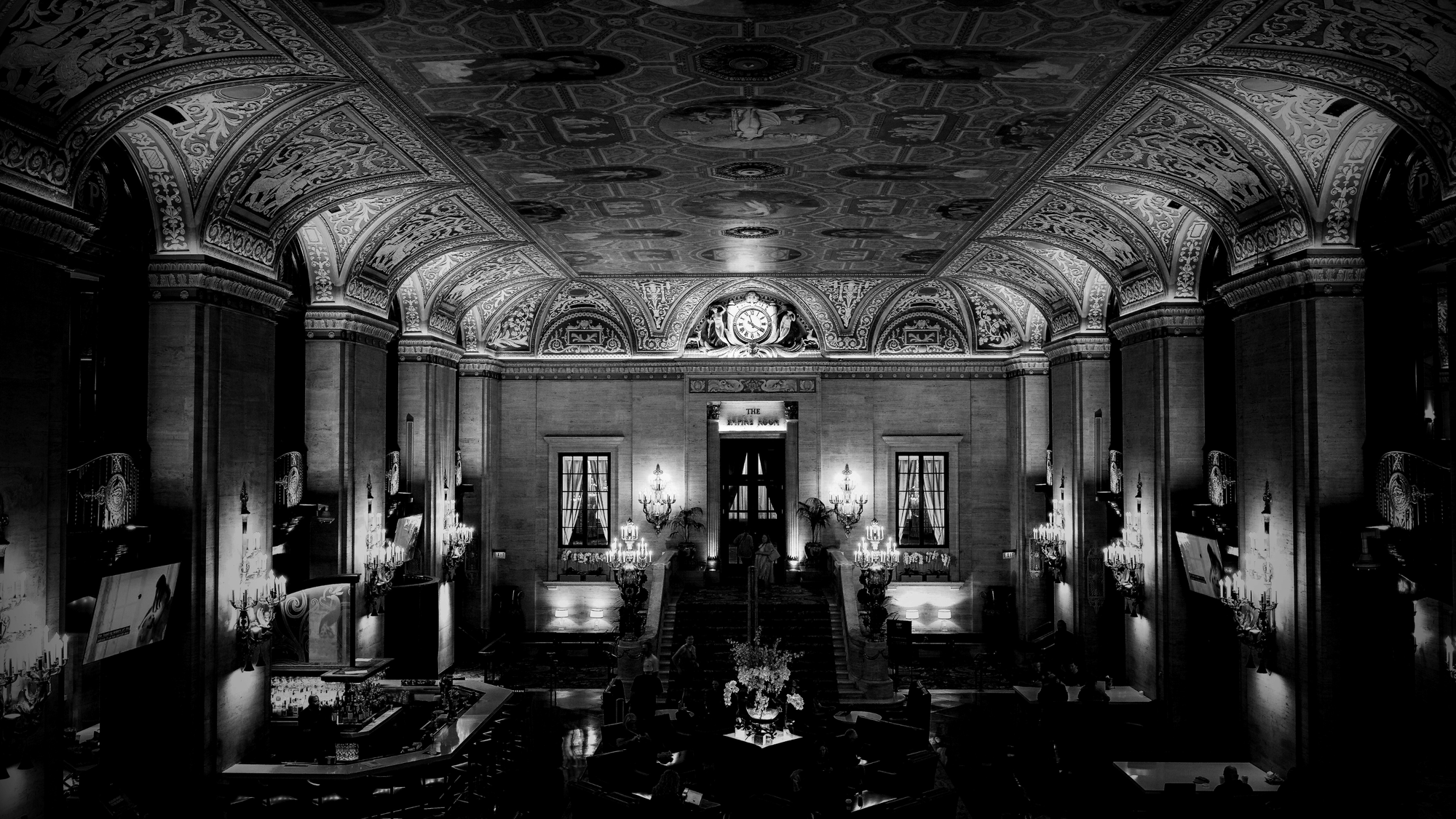 Black and white photograph of an ornate hotel restaurant lobby with symmetrical neoclassical architecture. Twin grand chandeliers illuminate the corridor flanked by tall fluted columns and elaborate arches. A clock is visible above the “Empire Room” entrance, with elegant sconces, draped windows, and a central floral arrangement.